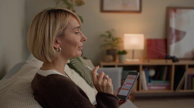 Handheld shot of focused adult Caucasian lady in casualwear sitting on sofa taking notes while checking menstrual period calendar on smartphone in living room