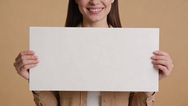 Smiling young woman holds a large blank white placard with both hands against a neutral beige background.