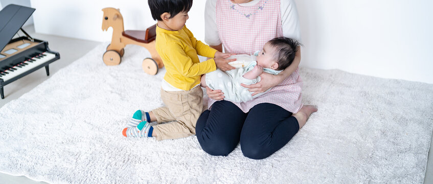 チェック柄のエプロンを着た女性が赤ちゃんを抱き、その隣で男の子が哺乳瓶を支えてミルクをあげるのを手伝っているカッ