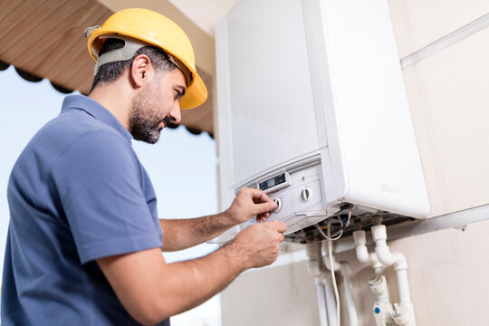 Professional handyman and electrician in blue overalls and yellow safety helmet performing home renovation, repairing boiler and installing electrical wiring in residential interior.