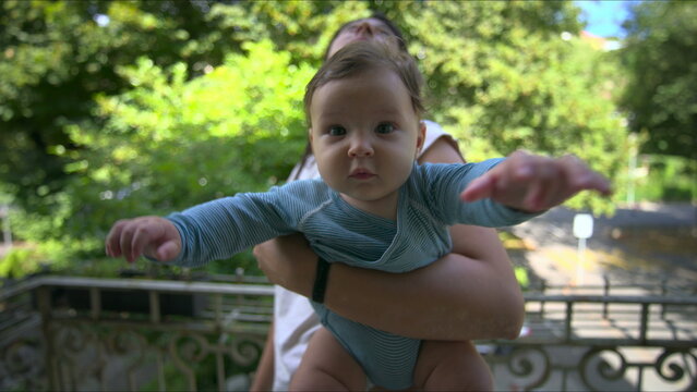 Mother standing on balcony holding baby, baby looking at camera while mother gazes into distance, peaceful family moment surrounded by greenery and decorative railing