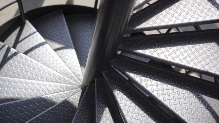 Metal spiral staircase with diamond plate treads descends around a central black pole in bright sunlight.
