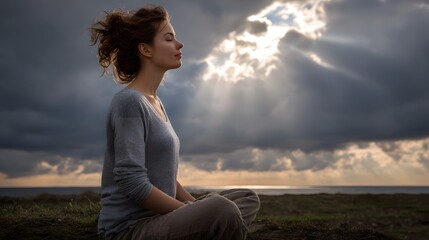 Young woman meditates peacefully outdoors under a dramatic sunlit sky overlooking the ocean