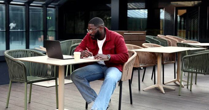 Young african american businessman sitting at a cafe, taking notes in his notebook while working remotely with his laptop. Focused professional enjoying coffee on an outdoor terrace