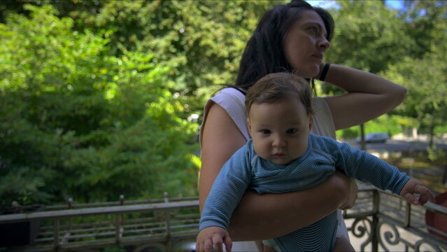 Mother embracing baby on balcony, baby resting calmly in arms, mother holding securely, lush green trees and residential area in background
