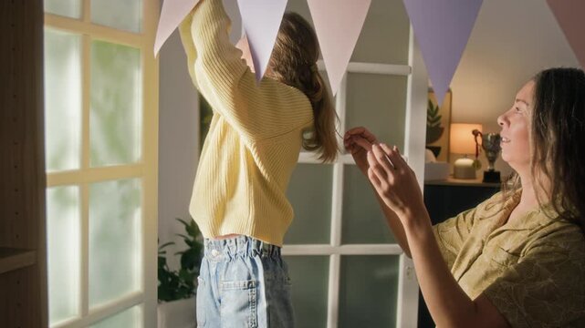 Positive gen alpha girl helping mother hang flag garland and they high-fiving each other while decorating living room