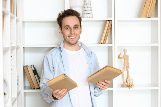 Smiling young man holding books in home library standing near bookshelf, casual student or reader portrait indoors with education, learning and reading concept