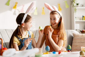Happy girls decorating Easter eggs wearing bunny ears, celebrating spring holiday