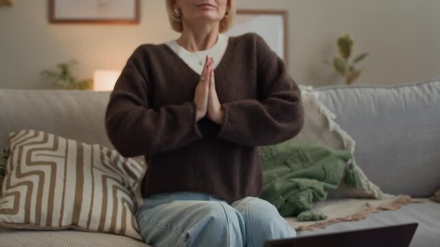 Tilt up shot of adult Caucasian woman starting meditation app on wireless laptop and sitting in prayer pose doing breathing exercise on couch in living room