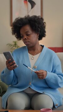 Vertical shot of young adult African American woman sitting on sofa checking result of pregnancy test and using mobile phone at home