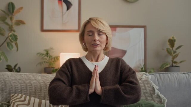 Zoom in shot of mindful Caucasian lady practicing prayer pose with hands by chest while meditating with application on wireless laptop on sofa in living room