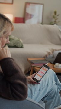 Vertical shot of focused adult Caucasian woman sitting on sofa using app on smartphone while tracking menstrual cycle in living room