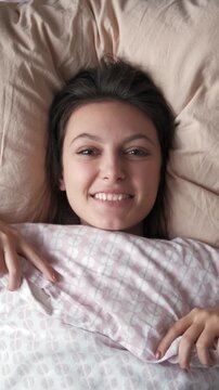 Young woman smiling and holding a soft duvet while lying in bed in the morning, embracing comfort and relaxation, enjoying a peaceful moment after waking up. Vertical shot