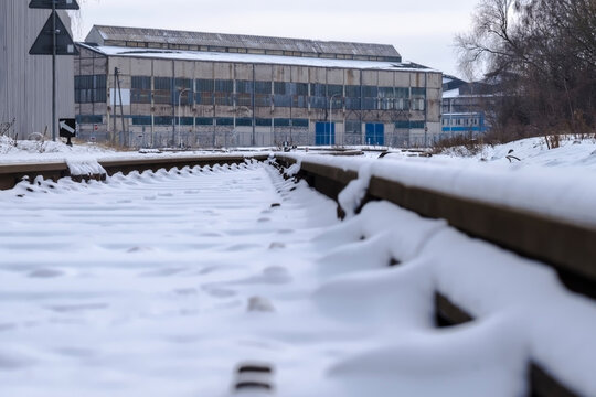 Tory kolejowe w śniegu przed fabryką. Pokryte śniegiem tory kolejowe prowadzą w kierunku fabryki przemysłowej w tle. Fabryka charakteryzuje się dużymi, wieloszybowymi oknami i fasadą z blachy.