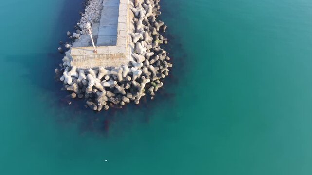 Aerial drone panorama of harbor pier and rocky breakwater stretching into clear turquoise sea water.