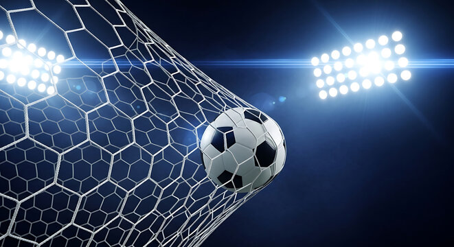 A soccer ball flies into the goal net under bright stadium lights during a nighttime match with a dark background