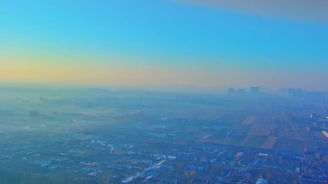 A peaceful drone shot capturing a traditional rural village covered in thick morning fog with distant city skyscrapers under a soft sunrise sky.