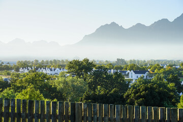 Obraz premium South Africa,Winery in Stellenbosch ,A serene suburban landscape with white houses nestled among green trees, framed by a wooden fence and distant misty mountains under a clear sky.