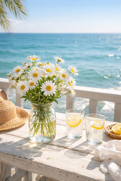 A bright seaside setting featuring fresh daisies in a glass vase and two glasses of lemon water on a white wooden table. With the turquoise ocean and palm leaves in the background, the scene...
