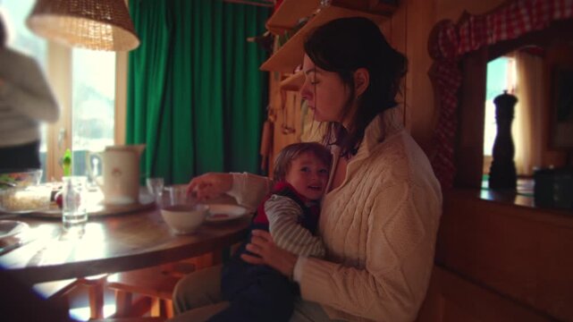 Mother eating meal while holding restless toddler in arms at wooden table inside cozy alpine chalet kitchen