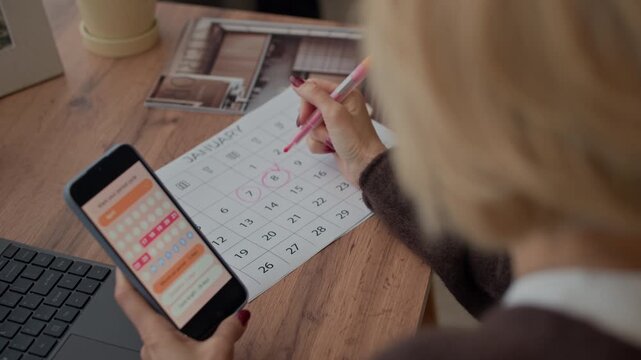 Over the shoulder view of adult blonde woman holding smartphone with menstrual period application while circling dates in calendar at home table