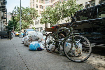 Obraz premium A green bicycle stands beside plastic garbage bags and parked vehicles on a downtown sidewalk. The scene shows everyday urban life, waste collection, and city transportation.