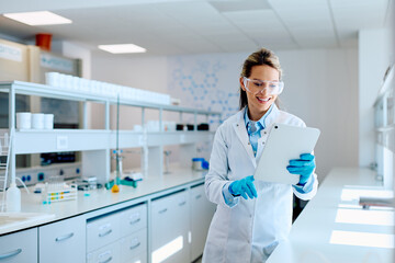 Naklejka premium Female scientist wearing safety glasses and lab coat, smiling while analyzing data on a tablet in a modern research laboratory