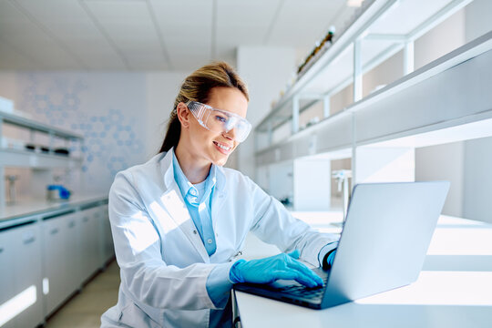 Young female scientist wearing lab coat, safety glasses, and gloves, working on a laptop in a modern laboratory