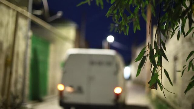 A van navigates a narrow, dimly lit street at night, with warm lights glowing from windows and reflections on wet pavement. captures urban life, delivery, and nighttime atmosphere.