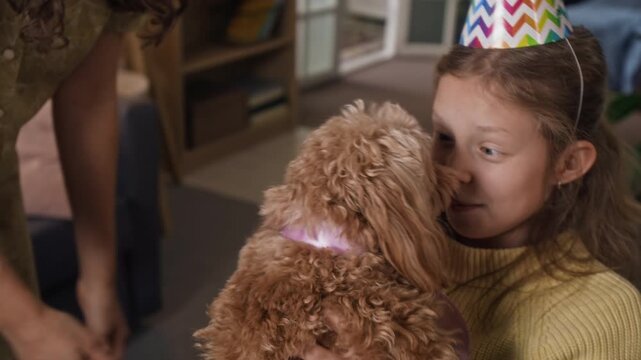 Long-haired woman giving cute moodle dog with pink ribbon to delighted gen alpha daughter celebrating birthday at home