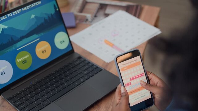 Cropped shot of African American woman checking menstrual cycle on smartphone while sitting in front of wireless laptop with meditation application at home table