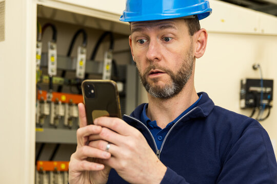Scared man, worker in helmet after seeing instructions on mobile phone. Maintenance, circuit breaker in electrical substation