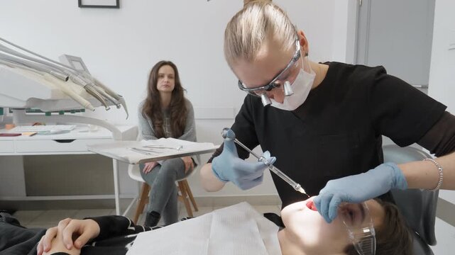 Dentist administers local anesthesia injection with syringe to patient in stomatology clinic chair while worried mother waits in background, illustrating dental phobia and fear of pain.