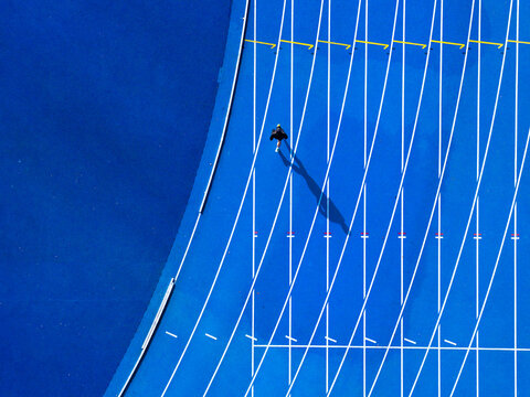 Aerial view of a lone runner casts a long shadow on the vibrant blue track, a striking contrast against the crisp white lines, Warsaw, Masovian Voivodeship, Poland.