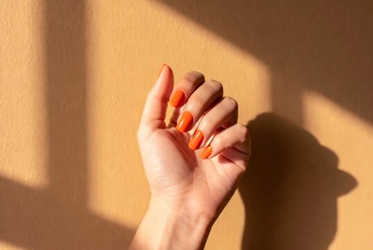 Subject: A hand with orange nails. Action: Holding up hands in shadow. Setting: A textured beige wall.