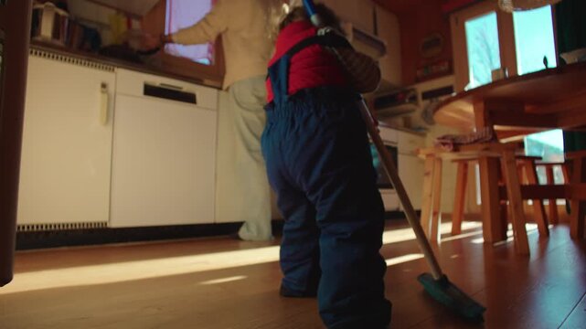 Small child mopping floor in kitchen while adult stands nearby, early responsibility and playful participation in home routine