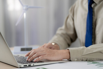 Business professional working on laptop at modern office with financial charts and wind turbine model, concept of renewable energy investment, sustainability strategy and green business technology.