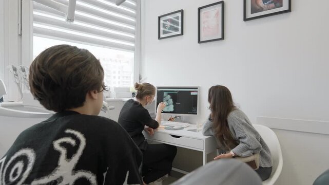 Stomatology doctor points at computer monitor screen explaining dental xray scan results to teenage boy patient and his mother, discussing oral cavity diagnosis and treatment plan.