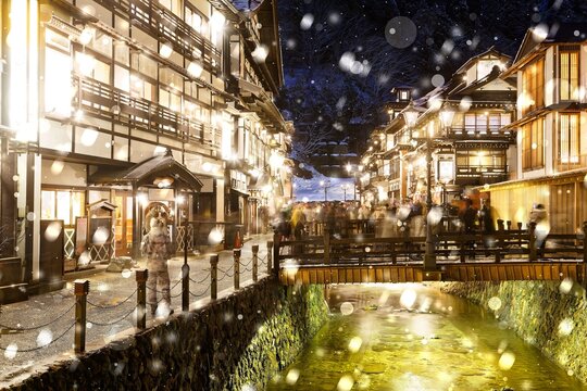 Night scenery of Ginzan Onsen, a famous hot spring town and tourist destination in Obanazawa, Yamagata, Japan, with bridges over a stream flanked by old wooden buildings on a cold snowy winter evening