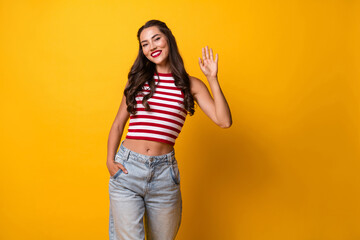 Young smiling woman in striped top and jeans waves hello against a bright yellow background