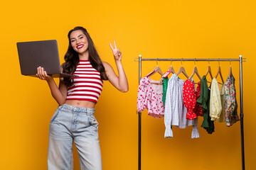 Young woman model with laptop and fashion rack smiling on bright yellow background