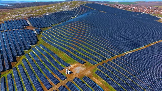 Vast solar panel array on rolling hills landscape. High angle view of rows of photovoltaic modules covering the terrain with a small town in the distance.
