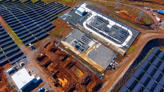 Aerial view of electrical substation at solar power plant. High angle shot of transformer station and power lines under construction next to solar panel rows.