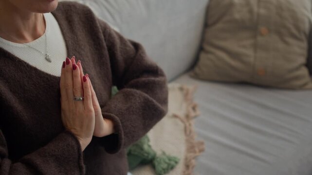 Handheld shot of relaxed middle-aged Caucasian woman in casualwear holding hands by chest while meditating with breathing exercise using application on wireless laptop at home