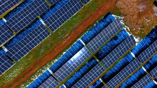 Diagonal aerial view of solar farm with sun glint. High angle perspective of solar panel arrays with bright sun reflection and patches of snow.
