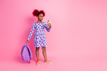 Young girl with pink hair wearing purple polka dot dress holds phone and backpack against pink...