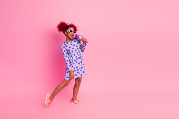 Young girl sings into pink microphone wearing a polka dot dress against a pink studio background in...