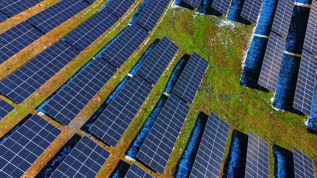 Aerial perspective of solar panels on green hillside. Diagonal rows of photovoltaic modules installed on a sloping terrain with grass and rocks.