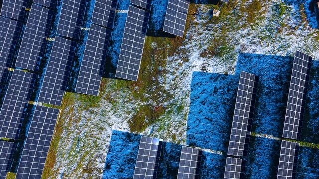 High angle shot of solar panels on snowy field. Photovoltaic modules with long shadows on a field partially covered with snow and grass.