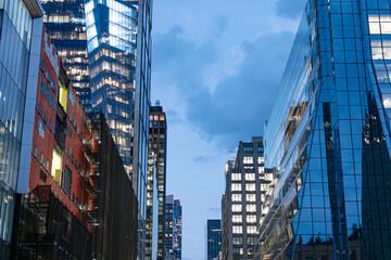 Obraz premium Glass skyscrapers rise into the blue evening sky in Hudson Yards, Midtown Manhattan. Illuminated office windows highlight business and corporate life in New York City.